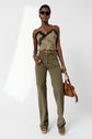 Woman wearing sunglasses, a floral lace-trim camisole, high-waisted olive patterned pantalon monogramme, platform shoes, and a brown belt. She holds a brown handbag and stands confidently against a plain white background.