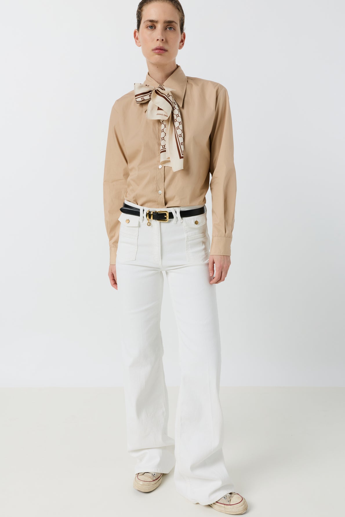 A person stands against a white background wearing a tan long-sleeve shirt with a patterned bow, coupe flare jean ANNA in a taille haute fit, a black belt, and beige sneakers.