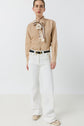 A person stands against a white background wearing a tan long-sleeve shirt with a patterned bow, coupe flare jean ANNA in a taille haute fit, a black belt, and beige sneakers.