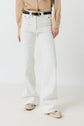 Person wearing a tan long-sleeve shirt, jean ANNA coupe flare blanc taille haute with gold buttons, a black belt, and worn white sneakers, standing against a plain white background.
