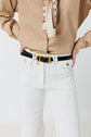 Person wearing a beige shirt, patterned scarf, black belt with gold buckle, and coupe flare jean ANNA taille haute in white, standing with hands on hips.