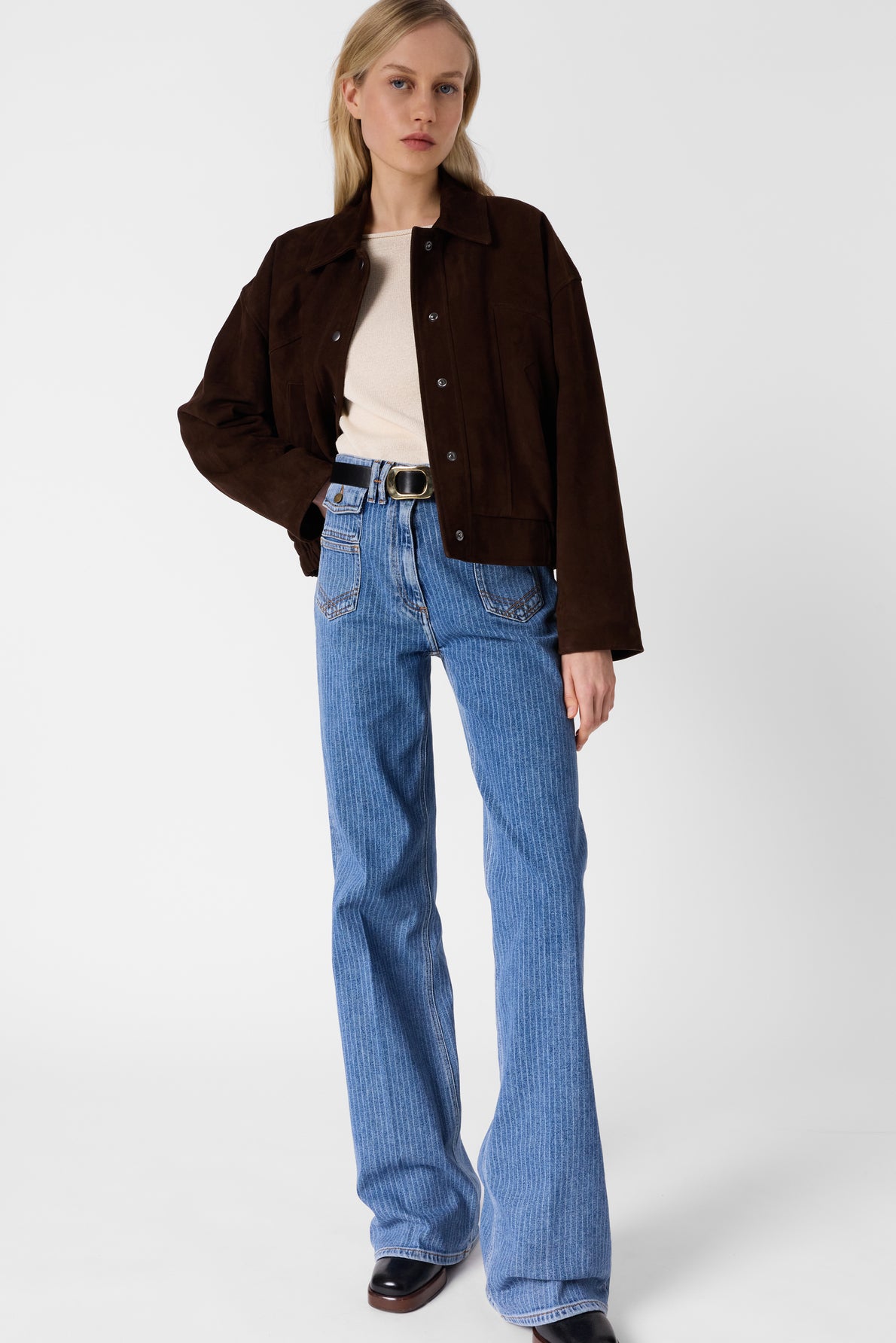A person stands against a white background wearing a dark brown jacket, off-white top, and a pair of blue jean ANNA taille haute à coupe flare. They have straight blond hair and a neutral expression, completed by black shoes.