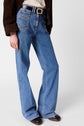 A person wearing blue pinstripe wide-leg jeans coupe flare jean ANNA with front pockets, a black belt with a gold buckle, a cream top, and a brown jacket stands against a plain white background.
