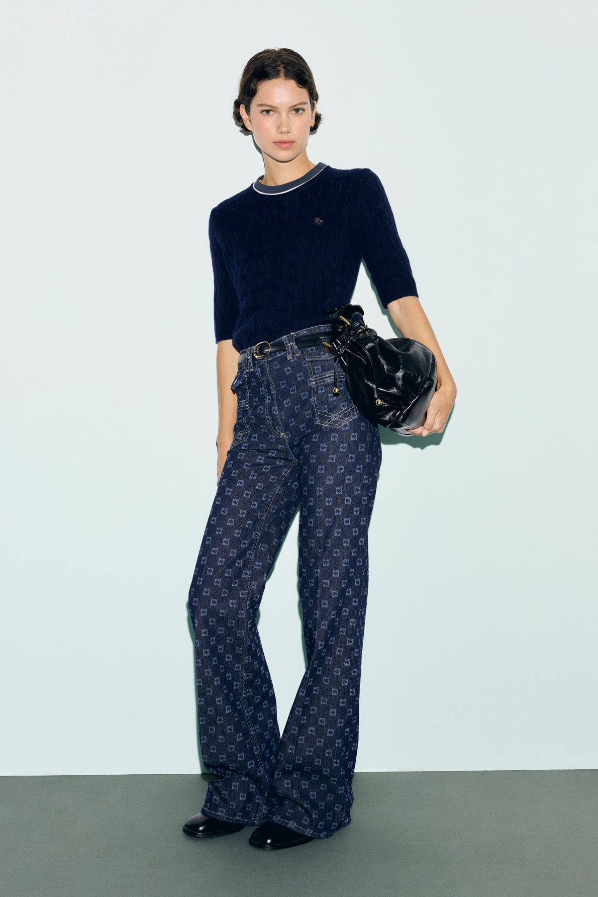 Woman stands against a light background wearing a navy short-sleeve sweater, high-waisted patterned jeans with a subtle motif monogramme, black belt, and shiny black bag, with short hair styled simply.