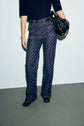 A person wearing dark blue patterned jean flare with a taille haute, a navy knit top, and black shoes holds a black handbag. The background is plain and light-colored.
