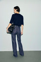 A person with short hair stands facing away, wearing a navy knit top and a pair of blue jeans taille haute à motif monogramme flare, holding a black bag on their left shoulder, against a plain light gray background.