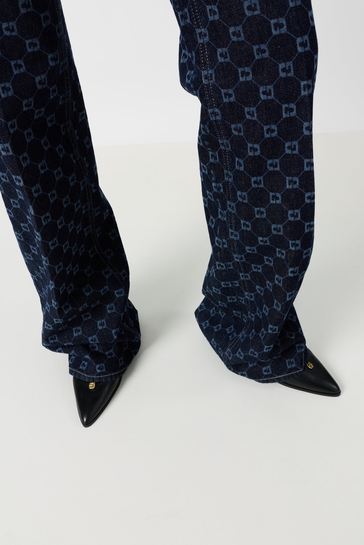 Person wearing dark blue patterned pants with a geometric motif monogramme and black pointed-toe shoes, standing on a light-colored floor.