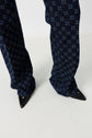 Person wearing dark blue patterned pants with a geometric motif monogramme and black pointed-toe shoes, standing on a light-colored floor.
