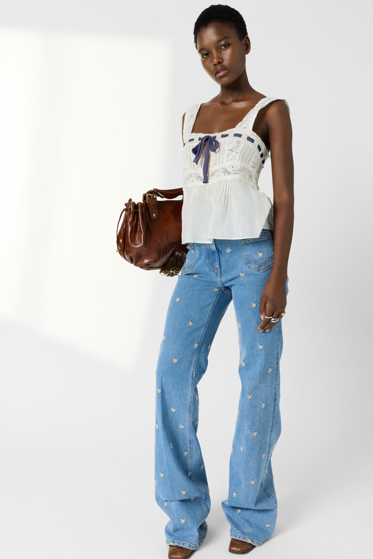 A woman wearing a white sleeveless top with bow detail, blue embroidered jeans, and brown heels stands holding a brown handbag, against a plain light background.