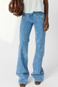 Person wearing light blue flared jeans with small white flower embroidery, a white top, brown shoes, and holding a large brown bag. Only the lower half of the body is visible.