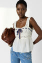 Woman wearing a white lace tank top with a navy ribbon, blue jeans with small embroidered flowers, and holding a brown leather bag, standing against a plain background.