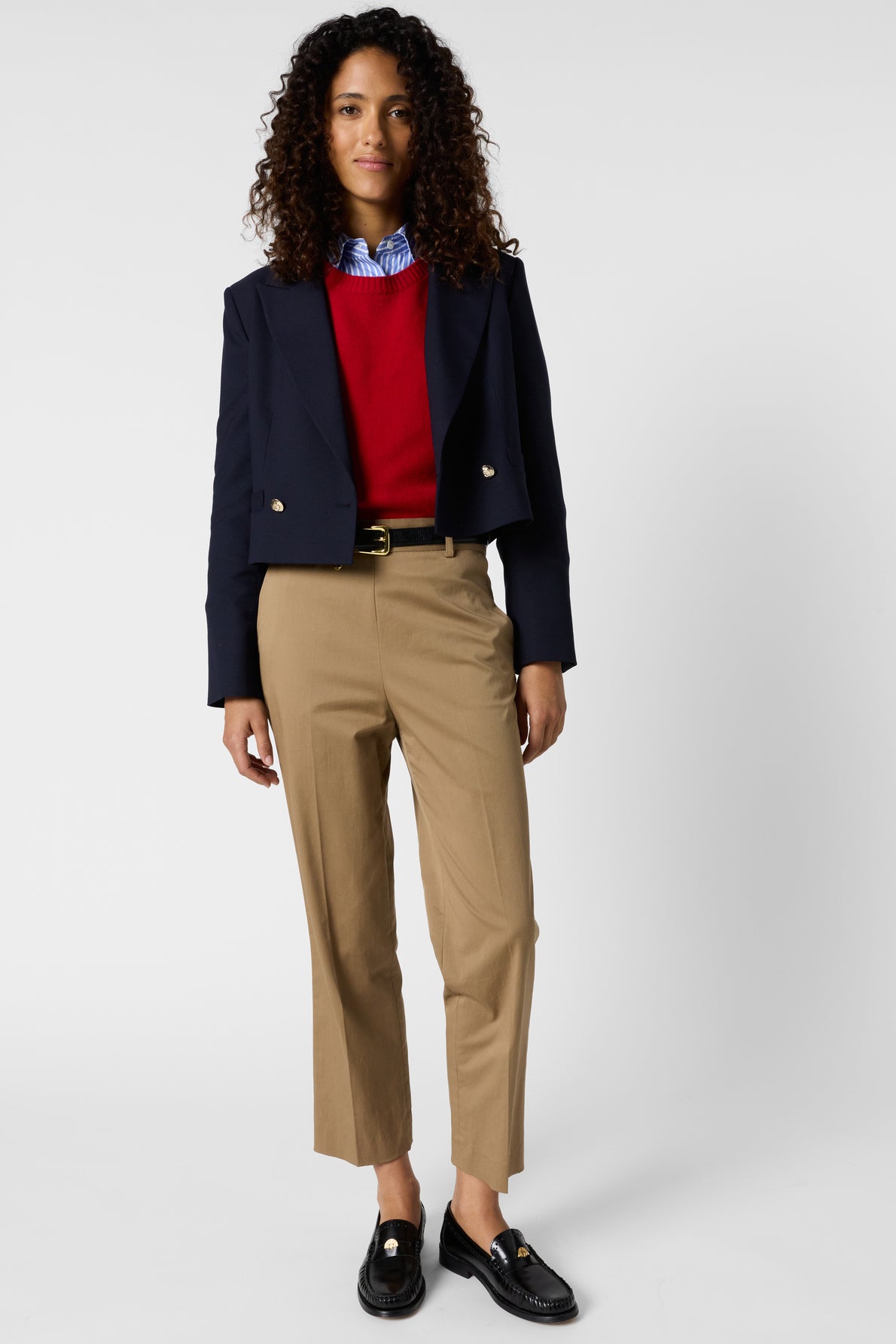 A woman with curly hair wears a navy blazer, red sweater, blue collared shirt, and a pantalon 7/8 coupe droite in tan cotton. She completes her look with black loafers and stands facing forward against a plain, light background.