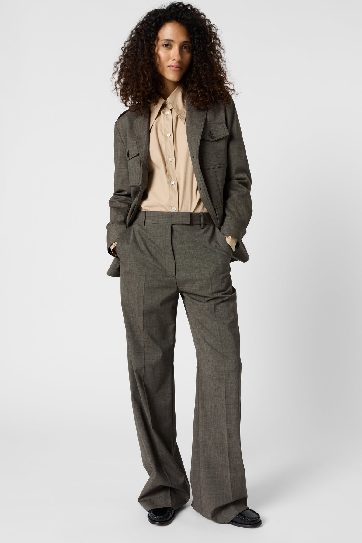 A person with curly hair stands facing forward, wearing a loose-fitting gray suit with un pantalon droit taille haute and a beige button-up shirt, hands in pockets, against a plain white background.