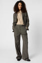A person with curly hair stands facing forward, wearing a loose-fitting gray suit with un pantalon droit taille haute and a beige button-up shirt, hands in pockets, against a plain white background.