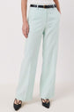 Person wearing pale mint green pantalon de tailleur in lin mélangé, a black belt, and black pointed-toe heels, with hands in their pockets, standing against a plain light background—an effortlessly chic tenue de bureau.