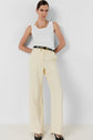 Woman poses in a white sleeveless top, high-waisted cream pantalon tailleur femme wide-leg pants, black belt with gold buckle, gold necklace, and black heels for a tenue bureau chic look against a plain white background.