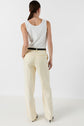 A woman stands facing away, wearing a white tank top and cream-colored high-waisted pantalon tailleur femme, paired with a black belt and black heels for a tenue bureau chic. Her hair is tied back in a low ponytail. The background is plain white.