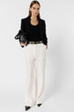 Woman wearing a black blazer, black top, white wide-leg pants with a belt, black pointed shoes, and holding a black handbag, standing against a plain white background.
