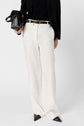 Person wearing white wide-leg trousers, a black top, black pointed shoes, a black blazer, and holding a black handbag. Outfit features a black belt with a gold buckle.