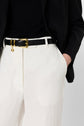 Person wearing white pants, a black belt with a gold buckle, a black top, and a black blazer. One hand is in their pocket. The outfit is stylish and contrasts dark and light colors.