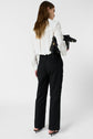 Woman standing with her back to the camera, wearing a white blouse and a pantalon noir coupe droite longueur 7/8, paired with heels and holding a black bag under her arm against a plain white background.