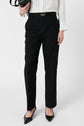 Person wearing black coupe droite tailored pants in a longueur 7/8 cut, a white long-sleeve blouse, and black pointed-toe shoes, holding a black handbag. The photo is cropped from shoulders down.