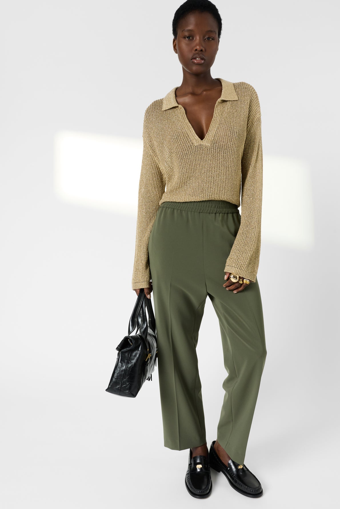 A person wearing a beige knit sweater with a deep V-neck, olive green pantalon fluide à taille élastiquée 7/8, and black loafers holds a black handbag, standing against a plain white background.