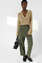 A person wearing a beige knit sweater with a deep V-neck, olive green pantalon fluide à taille élastiquée 7/8, and black loafers holds a black handbag, standing against a plain white background.
