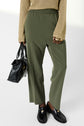 A person wearing an oversized beige sweater, a pantalon 7/8 olive green à taille élastiquée, and black loafers holds a black handbag. Only the lower body is visible against a plain background.
