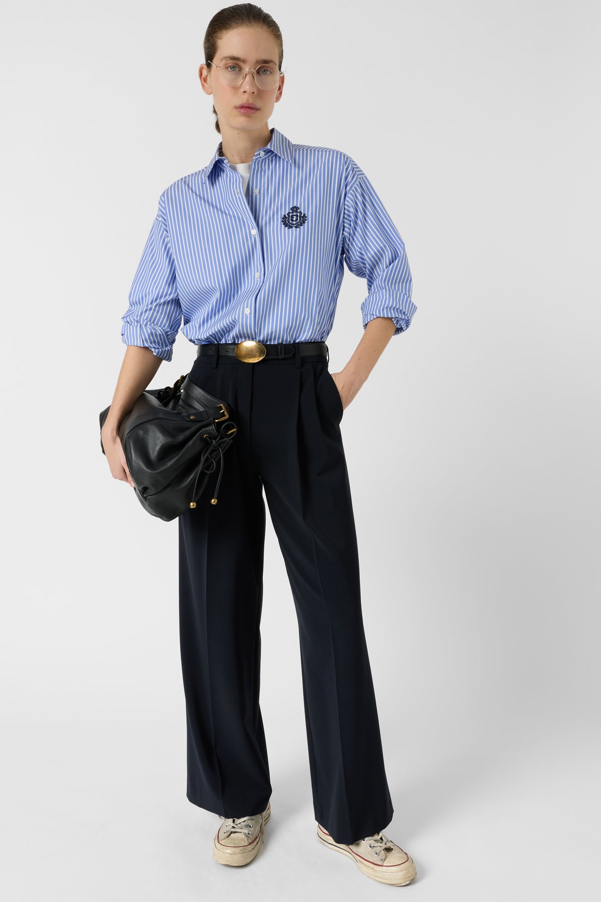 Person standing against a white background, wearing a blue striped shirt with rolled-up sleeves, navy wide-leg pants, beige sneakers, and holding a black handbag under one arm.