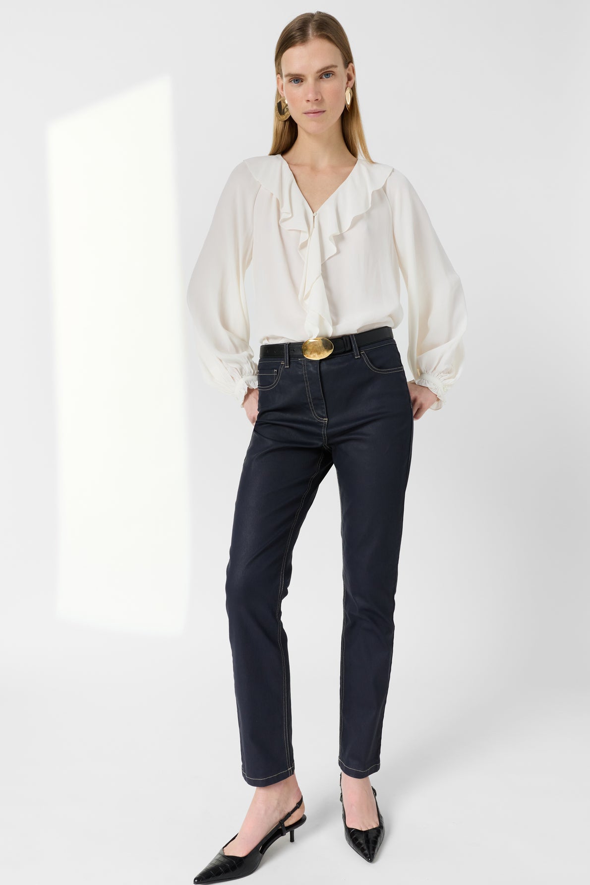 Woman standing against a white background, wearing a white ruffled blouse and a jean slim bleu marine à coupe ajustée with a large belt buckle, black pointed shoes, and gold earrings. She has straight hair and a neutral expression.