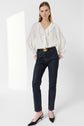Woman standing against a white background, wearing a white ruffled blouse and a jean slim bleu marine à coupe ajustée with a large belt buckle, black pointed shoes, and gold earrings. She has straight hair and a neutral expression.