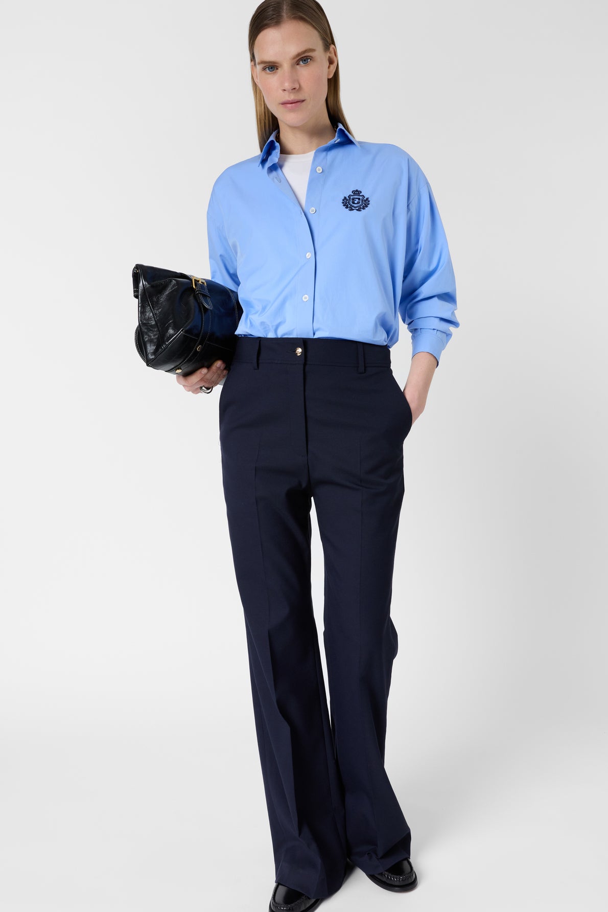A woman stands against a plain background wearing a light blue button-up shirt, dark wide-leg trousers, and black shoes. She holds a black clutch bag and has straight, light brown hair.