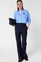 A woman stands against a plain background wearing a light blue button-up shirt, dark wide-leg trousers, and black shoes. She holds a black clutch bag and has straight, light brown hair.