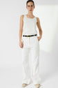 A person stands against a white background wearing a sleeveless white top, white wide-leg pants, a black belt with a gold buckle, and beige shoes. Their hair is pulled back.