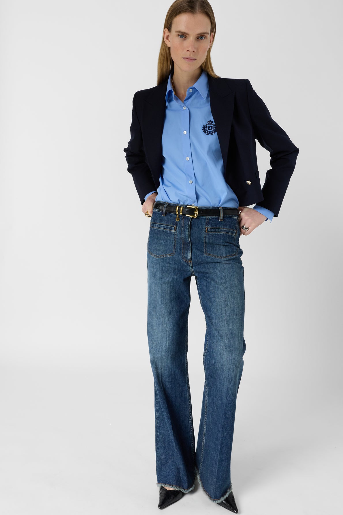A person stands facing forward, wearing a dark blazer over a blue button-up shirt, blue jeans, a black belt, and pointed black shoes. Their hands are in their jeans pockets.