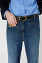 A person wearing blue jeans, a blue shirt, and a black belt with a gold buckle stands with one hand in their pocket, displaying gold rings.