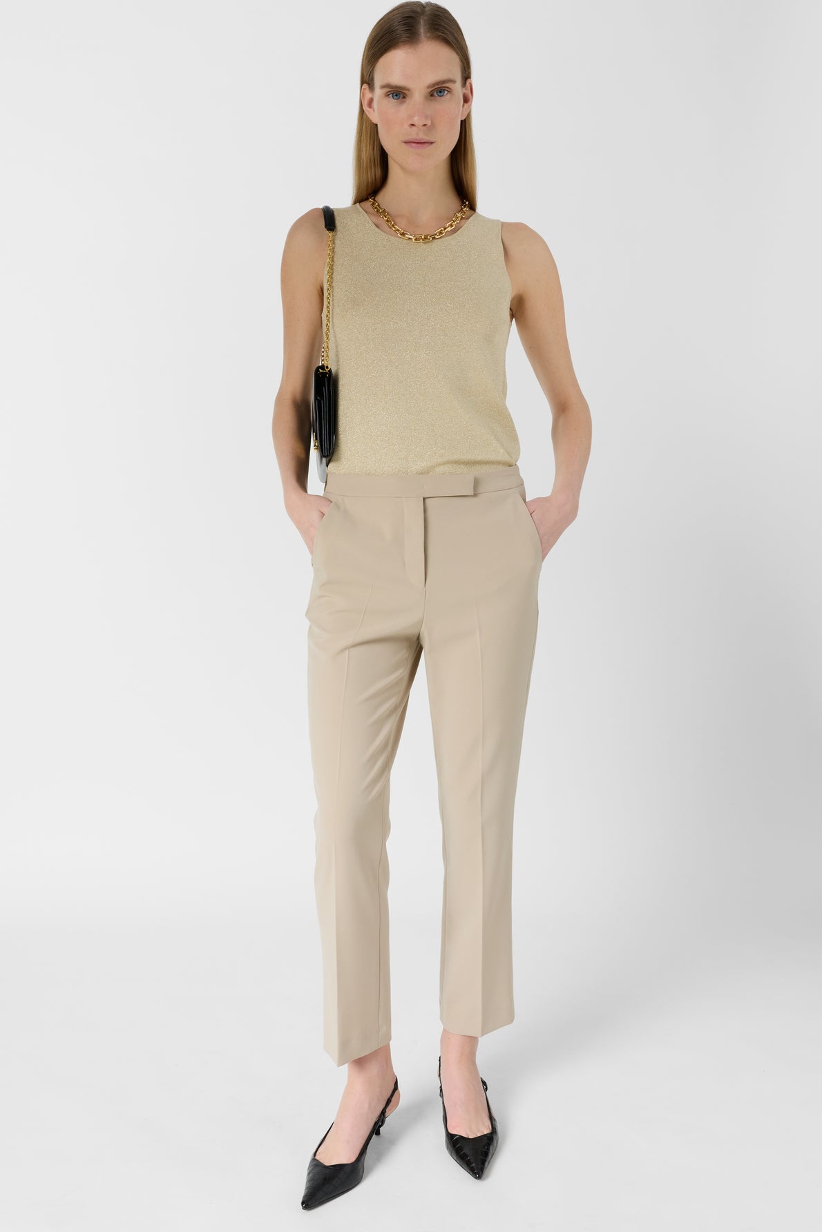 A woman stands against a white background wearing a gold sleeveless top, a chic pantalon 7/8 beige de tailleur, black pointed heels, a gold necklace, and a black shoulder bag. Her hands are in her pockets, and she has straight, loose hair.