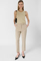 A woman stands against a white background wearing a gold sleeveless top, a chic pantalon 7/8 beige de tailleur, black pointed heels, a gold necklace, and a black shoulder bag. Her hands are in her pockets, and she has straight, loose hair.