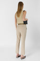 A woman with straight, light brown hair wears a sleeveless beige top, matching pantalon de tailleur beige 7/8, black sandals, and carries a small black purse with a gold chain. She stands against a white background, facing away from the camera.