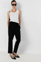 Woman wearing black sunglasses, a white sleeveless top, a chic pantalon tailleur femme noir 7/8, black pointed-toe heels, and a gold chain necklace, standing against a plain white wall with one hand in her pocket.