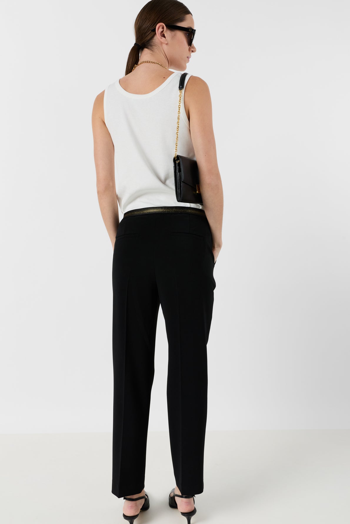 A person wearing a white sleeveless top, a chic pantalon tailleur femme noir, black sandals, sunglasses, and a black shoulder bag with a gold chain stands facing away against a plain white background.