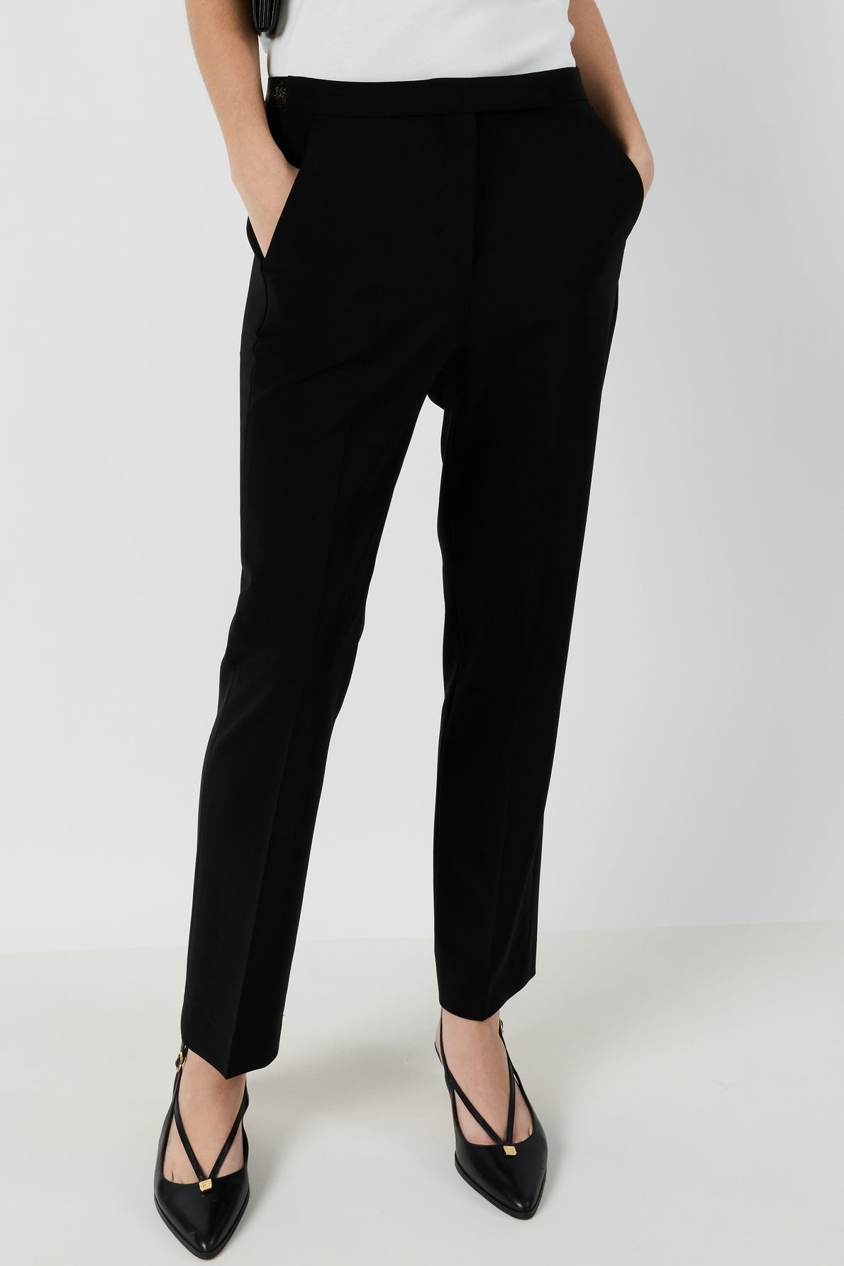 Person wearing a pantalon tailleur femme noir chic, tailored in a 7/8 cut, paired with black pointed-toe heels featuring gold tips, standing against a plain light background with hands in pockets and upper body mostly out of frame.