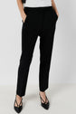 Person wearing a pantalon tailleur femme noir chic, tailored in a 7/8 cut, paired with black pointed-toe heels featuring gold tips, standing against a plain light background with hands in pockets and upper body mostly out of frame.