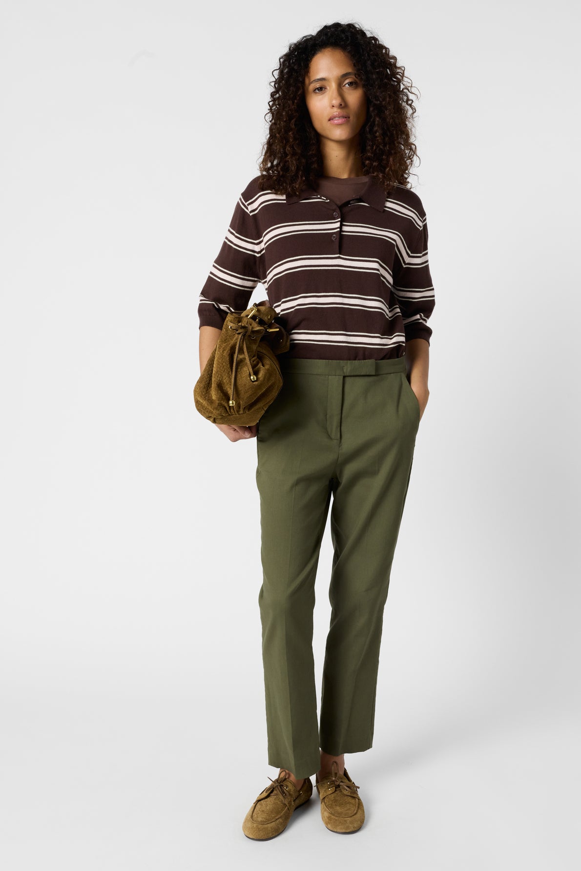 Woman standing against a white background, wearing a brown and white striped top, olive green pants, and brown shoes. She holds a brown suede drawstring bag and has curly hair.