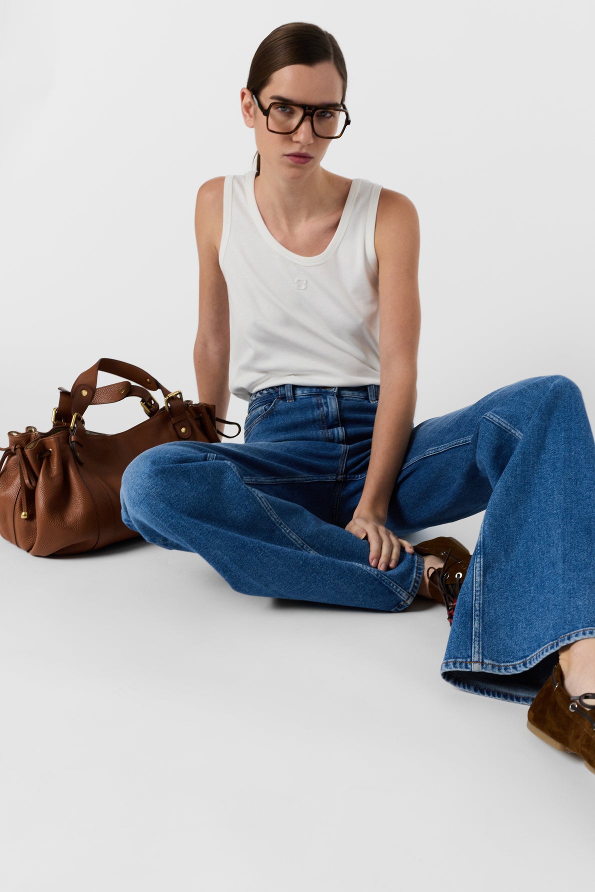 Woman with brown hair and glasses wears a white sleeveless top and blue jeans, sitting on the floor with legs crossed. A brown handbag and brown shoes are next to her against a plain white background.