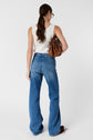 A woman with long brown hair in a ponytail wears a white sleeveless top and blue jeans, holding a brown handbag over her shoulder, standing against a plain white background.