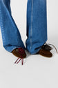 Close-up of a person wearing wide-leg blue jeans and brown suede loafers. The left shoe features a red ribbon bow decoration, while the right shoe features a decorative pinecone ornament.