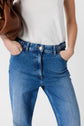 A person wearing blue jeans and a white shirt stands with hands in pockets, holding a brown bag under one arm. Only the torso and upper legs are visible.