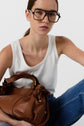 A woman wearing large glasses, a white sleeveless top, and blue jeans holds a brown leather handbag, sitting against a plain white background.